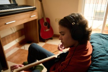 Bir genç gitar çalar, kulaklık takar, rahat bir odada pratik yapar. Yakınlarda bir kırmızı gitar ve klavye odaklanmış bir müzik oturumu oluşturur.