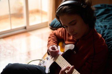 Genç bir çocuk kanepeye oturur, kulaklık takar ve elektro gitar çalar. Sıcak bir ışık ve rahat bir ruh hali evde kişisel müzik hobisinin altını çizer.