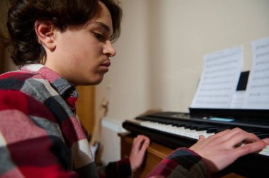 Ortadoğu kökenli genç bir çocuk bir piyanonun başına oturmuş, notalara odaklanmış. Konforlu ev dekorasyonu pratik, adanmışlık ve müzik yapmanın sessiz neşesini yakalıyor..