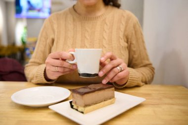 Samimi bir kafe sahnesinde bej kazaklı bir kadın elinde beyaz bir kahve fincanı, tabağında çikolatalı kek ve sıcak ahşap bir masa tutuyordu..