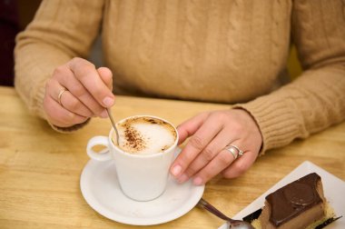 Sıcak bir kafe anı, bej kazaklı, köpüklü bir kapuçino karıştıran ve bardağın yanındaki tabakta çikolatalı kek dilimi olan birini gösterir..