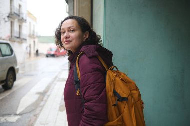 Yanmış turuncu sırt çantalı kendine güvenen bir kadın şehrin kaldırımında duruyor ve nazikçe gülümsüyor. Şehir sahnesinde ıslak kaldırım ve ışık görünüyor. Bu da sıradan bir seyahat anı olduğunu gösteriyor..