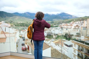 Bordo ceketli bir kadın, turuncu bir sırt çantasıyla ıslak bir kasabaya camdan bir balkondan bakıyor. Yağmurlu bir günde, uzaktaki dağlara bakıyor..