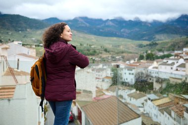 Kestane renkli balkon ceketli bir kadın cam bir balkonda duruyor ve camında yağmur yağan bir vadi kasabasına bakıyor. Uzakta dağlar belirir, sakin ve yansıtıcı bir yolculuk anı yaratır..