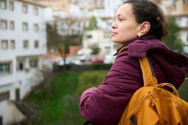 Kestane rengi ceketli ve hardal çantalı bir kadın bulanık bir şehir manzarasına bakıyor. Şehir ortamında huzur, yansıma ve seyahat anları yaşıyor..