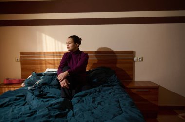 Contemplative woman sits on a bed in a dim bedroom, wrapped in a blue blanket. Warm light casts soft shadows, creating a cozy, intimate mood.