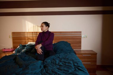 Dalgın bir kadın, sıcacık, güneşli bir yatak odasında, düzgün bir şekilde yapılmış bir yatağa oturmuş, üzerinde mor bir balıkçı yaka var. Mavi yorgan ve ahşap mobilyalar sakin, samimi bir ev sahnesi yaratır..