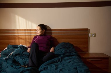 Bir kadın, yumuşak ışıklı bir yatak odasında, bir yatakta, tahta bir yatak başlığına yaslanmış olarak oturuyor. Mor bir bluz içinde, mavi bir yorgan ve sıcak bir sabah ışığı ile rahatlıyor..