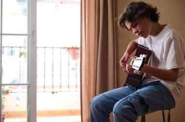 Genç çocuk akustik gitarını güneşli bir balkon penceresinde çalışıyor, akorlara odaklanıyor ve rahat ev müziği çalışmalarının ve gençlik yaratıcılığının tadını çıkarıyor..
