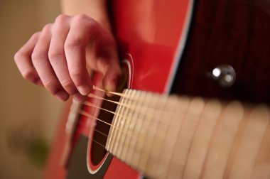 Kırmızı akustik gitarın tellerini koparan, parmaklar, teller ve ses panosuna odaklanan sığ, sığ ve samimi bir müzik ortamı olan bir elin yakın çekimi..