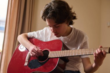 Teen focused on playing a red acoustic guitar indoors, practicing chords and learning music in a casual home setting with natural light.