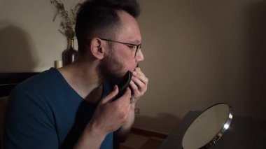 Bir adam sakalını elektrikli traş makinesiyle titizlikle keser, evde aynanın önünde kişisel bakım rutinine odaklanır..