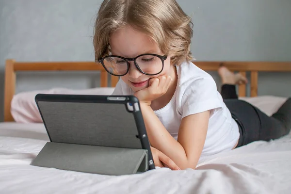 Mała dziewczynka w okularach odrabia pracę domową online, leżąc na łóżku w domu. Komunikacja z krewnymi online. Koncepcja początku nowego roku szkolnego — Zdjęcie stockowe