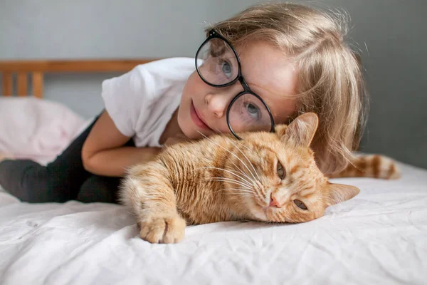 Holčička s brýlemi leží na posteli a objímá tlustou zrzavou kočku. — Stock fotografie