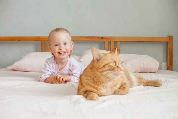 Ein kleines süßes Mädchen sitzt mit einer dicken Ingwerkatze auf dem Bett, streicht und spielt mit ihm. — Stockfoto