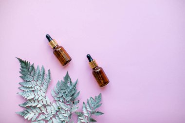 Kozmetik serum şişelerinin ve pembe arka plandaki dekoratif eğrelti otu dalının düzeni.