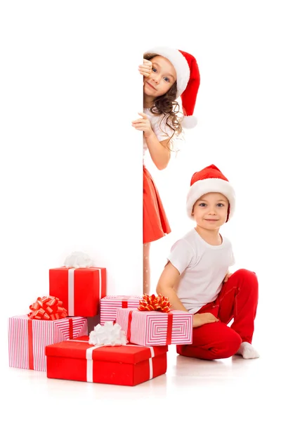Glückliche kleine Kinder mit Weihnachtsmannmütze, die hinter einer leeren Werbetafel hervorlugen — Stockfoto