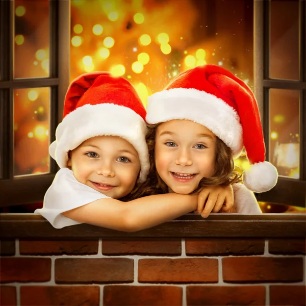 Niños felices en Santa Sombrero miran por la ventana en Navidad —  Fotos de Stock