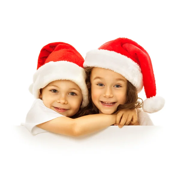Ευτυχισμένος παιδάκια στα Santa καπέλο που κρυφοκοιτάζει από πίσω από κενό σημάδι πινακίδα — Φωτογραφία Αρχείου