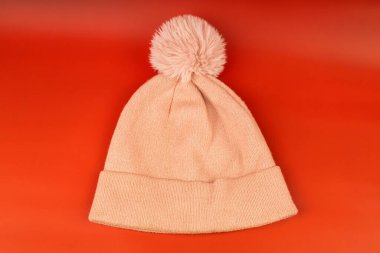 Yumuşak yünden yapılmış, büyük pofuduk bir ponponu olan zarif, pembe bir kış beresi. Şapka, dokusunu ve rengini vurgulayan canlı kırmızı bir yüzey üzerine kuruludur. Kış için klasik bir aksesuar