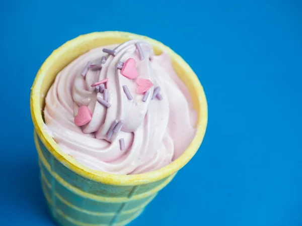 Pembe yumuşak dondurma, pembe kalpler serpiştirilmiş. Sarı bir waffle bardağında, mavi arka planda, en üstte..