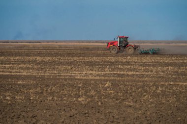 Mavi sabanı olan büyük kırmızı bir traktör mahsul ekmeden önce toprağı sürer. Mavi gökyüzünün arka planına karşı. Aşağıda bir yazıt yeri var..