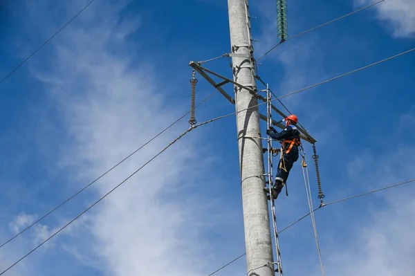 Turuncu kasklı bir elektrikçi mavi gökyüzüne karşı yüksek voltajlı bir güç kulesine tırmanıyor..