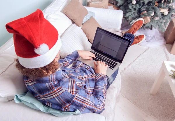 Jonge man met laptop op een bank in de buurt van een kerstboom. — Stockfoto