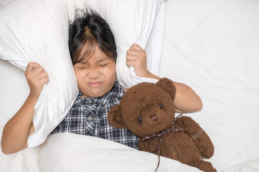 Little girl lying in bed covering head with pillow because too loud annoying noise. Irritated child suffering from noisy neighbors, trying to sleep after alarm wake-up signal