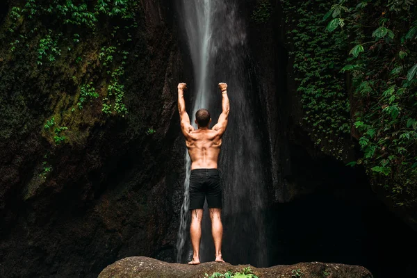 Back view of a man standing at a waterfall with his hands raised. The man at the waterfall raises his hands up. Man at the waterfall. Travel to Bali. Man on the background of nature. Copy space