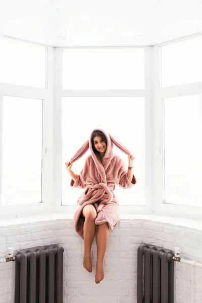 A beautiful woman in a dressing gown is sitting on the windowsill. A woman in a bunny costume. The girl at the window. A pretty girl in a pink robe.