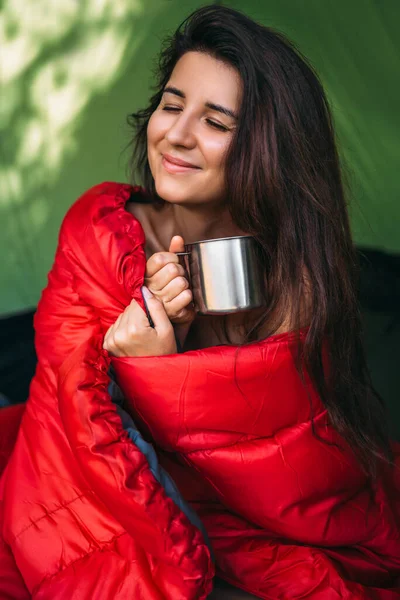 A happy tourist woman is sitting in a tent and drinking tea. Camp in the tent - young girl on the camping. A female tourist in a sleeping bag, drinking hot tea. Morning at the campsite. Portrait