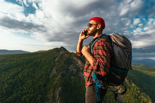 リュックを背負った赤い帽子の男性旅行者が山を背景に電話で話している ハイキング用の服を着た男が山の日没時にスマートフォンで話している 電話での会話 — ストック写真