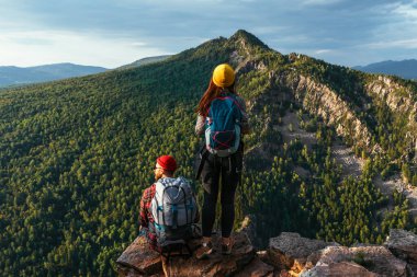 A man and a woman with backpacks on the mountain admire the panoramic view. A couple in love on a rock admires the beautiful views. Travelers in the mountains at sunset. A couple on a hike. Copy space