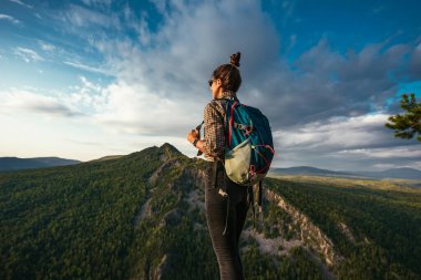 Sırt çantalı genç bir turist kız dağın tepesinden gün batımının tadını çıkarıyor. Dağların arka planında bir gezgin. Sırt çantalı bir turist dağın tepesinde duruyor.