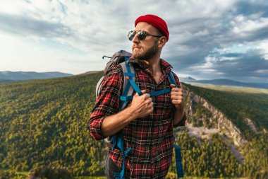 Dağın tepesinde sırt çantası olan sakallı bir gezgin. Kırmızı şapkalı ve güneş gözlüklü bir gezginin portresi. Sırt çantalı bir turist bir dağın arka planında duruyor. Boşluğu kopyala