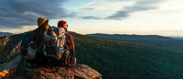 这对夫妇在山上遇见了日落 两个旅行者坐在悬崖边上欣赏美丽的全景 在日落时分背着背包在山上旅行的人 — 图库照片