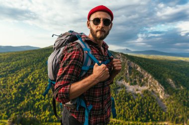 Dağın tepesinde sırt çantası olan sakallı bir gezgin. Kırmızı şapkalı ve güneş gözlüklü bir gezginin portresi. Sırt çantalı bir turist bir dağın arka planında duruyor. Boşluğu kopyala
