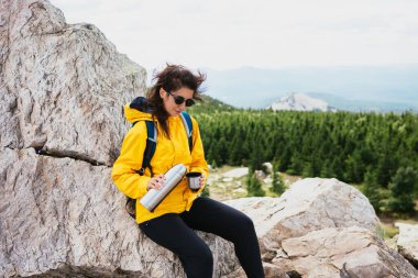 Bir turist kız dağa tırmandıktan sonra dinleniyor. Mutlu bir turist yeşil bir ormandaki termostan kahve içer. Dişi bir gezgin dağlardaki termostan sıcak çay içer..