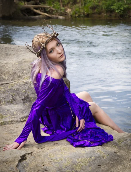 Νεαρή γυναίκα σε μωβ φόρεμα κάθεται από ένα ποτάμι — Φωτογραφία Αρχείου