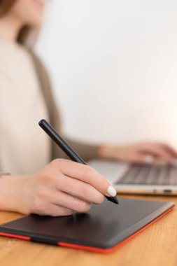 Grafik tasarımcısı dijital tablet ve dizüstü bilgisayarla çalışıyor. Ahşap çalışma alanı. Ev ofisinden bağımsız çalışan bir kadın. Elinde kalem, arka planında bulanıklık olan seçici odaklanma. Fotoğrafı kapat..