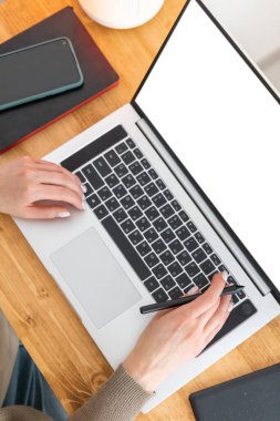 Beyaz kadın ahşap masada oturuyor ve dizüstü bilgisayarın başında çalışıyor. Kadın elinde defterle boş ekranda kitap okuyor. Ev ofisinden çalışan serbest çalışan biri. Üst manzara, düz uzanma.