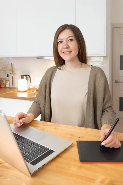 Smiling caucasian woman graphic designer sitting at workspace and looking into camera. Girl in beige t-shirt and grey cardigan work from home office. On the wooden desk laptop and digital tablet.