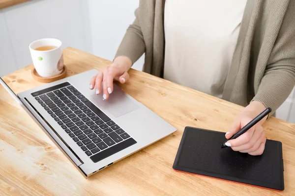 Açık tişörtlü, gri hırkalı, resimlerini çizen ya da resimlerini çizen tanınmamış bir kadın. Laptop, bir fincan çay ve tablet tahta masada. Evden uzakta çalışan genç bir serbest yazar..
