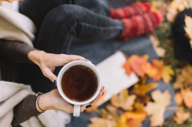 Beyaz kadın elinde siyah çayla beyaz bardak tutuyor. Ormanda sonbahar pikniği. Sıcak sıcak içecek. Seçici odaklanma. Kitap ve turuncu akçaağaç yaprakları arka planda. Doğada zaman geçirmek. Düz yatıyordu.