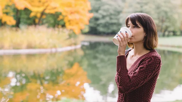 Attractive woman in purple sweater drinking coffee from a white mock up cup. Lake and forest on blurred background. Hot beverage for cold days. Changing of seasons concept.
