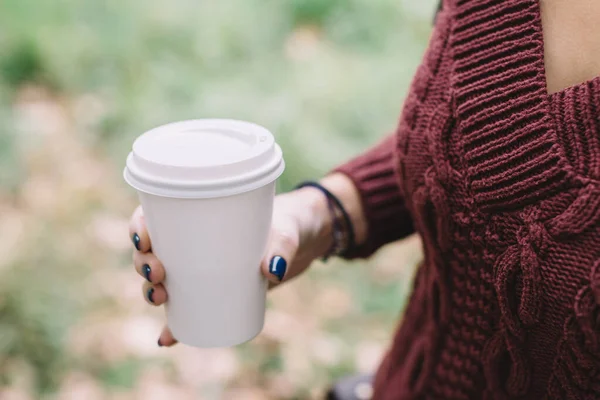 Unrecognizable girl in red sweater holding white blank paper cup with coffee or tea. Hot beverage for warming on cold days. Changing of seasons concept. Soft focus. Takeaway latte or cappuccino.