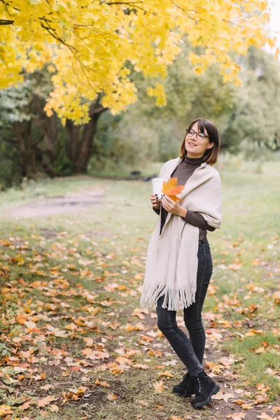 Happy girl in black jeans, brown sweater and beige scarf holding white paper cup with coffee or tea. Takeaway latte or cappuccino. Hot beverage for cold days. Walking in autumn forest.
