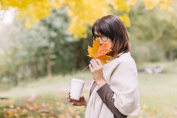 ガラスのシャイなブルネットの女の子は 紅茶やコーヒーを片手に白いモックアップカップを手に持ち オレンジメープルリーフの後ろに隠れています 温かい飲み物 秋のシーズン 背景がぼやけている テイクアウトドリンク — ストック写真