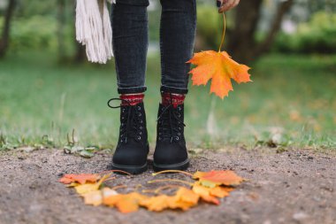 Gri kotlu, kırmızı örgü çoraplı, bej eşarplı ve elinde turuncu akçaağaç yaprağı tutan siyah çizmeli şık bir beyaz kadın. Sarı yapraklar kızdan önce gelir. Arka planda yeşil çimen. Sezon konsepti değişikliği.